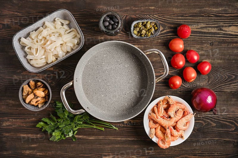 Fresh seafood ingredients are arranged on a wooden kitchen table. There are shrimp, mussels, squid, tomatoes, onions, olives, and capers. An empty pot is placed in the center ready for cooking
