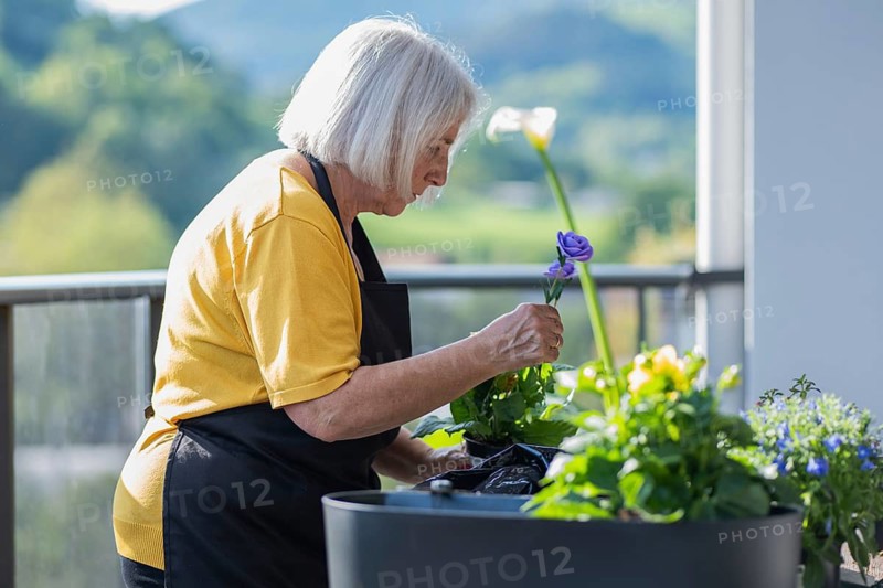Elderly woman in a yellow shirt and black apron tending to a variety of colorful flowers on an outdoor deck, symbolizing tranquility and love for gardening