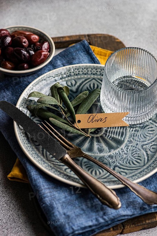 A tasteful arrangement of Greek BBQ olives on a ceramic plate, accompanied by a glass, rustic utensils, and textured linens. The scene captures a balance of elegance and tradition, perfect for culinary themes