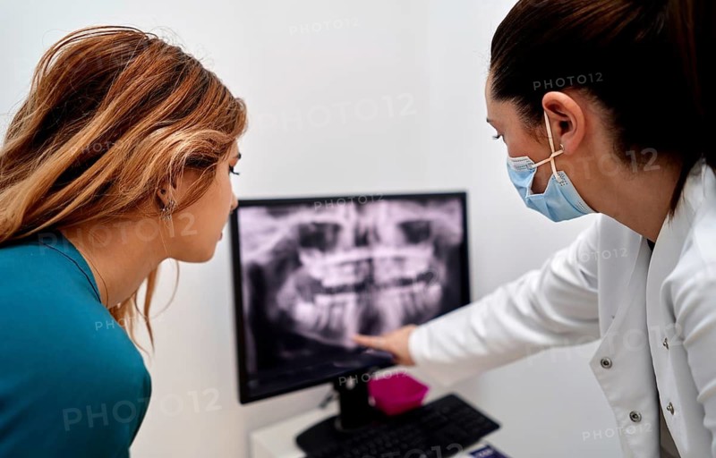 A dentist in a dental clinic shows an x-ray on a computer screen to a patient, explaining details while both focus on the image. The patient listens attentively to the dentist