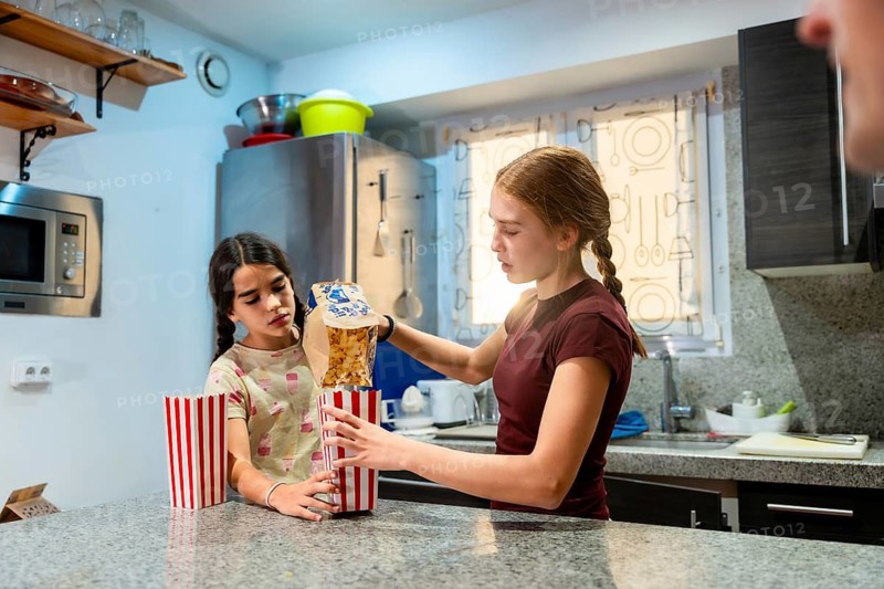 Two young sisters standing in a kitchen, one pouring popcorn from a bag into striped paper containers, preparing for an evening of entertainment together at home