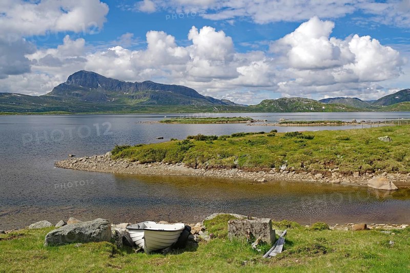 Lake with a small boat on the shore surrounded by green mountains under blue sky, Bygdin, Jotunheimvegen, Mjolkevegen, Valdesflye, Norway