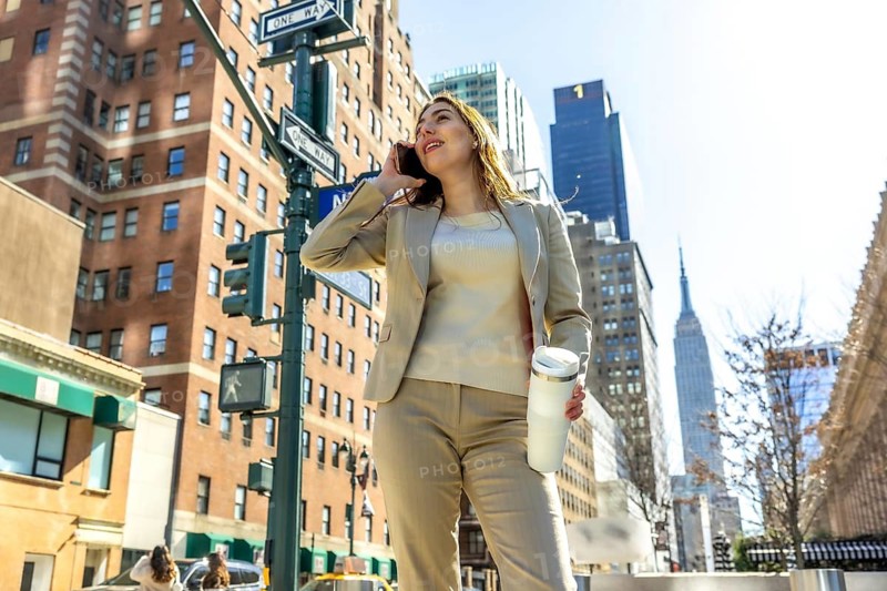 Professional businesswoman enjoying a call on her mobile phone while holding a travel mug, walking confidently along a sunny manhattan street with iconic architecture in the background