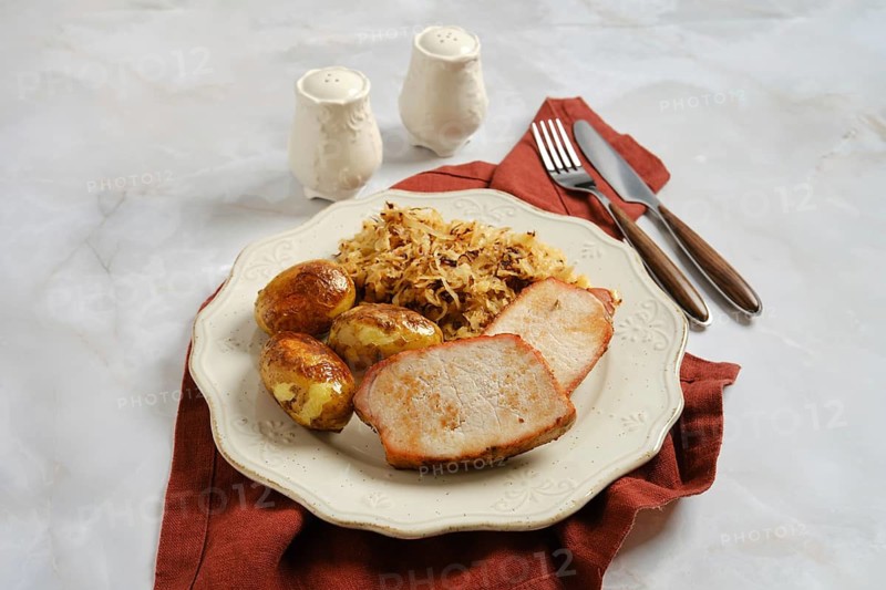 Plate with sliced roasted pork chops, roasted baby potatoes, and fried sauerkraut on table. Set includes two salt shakers and utensils showing mealtime setting