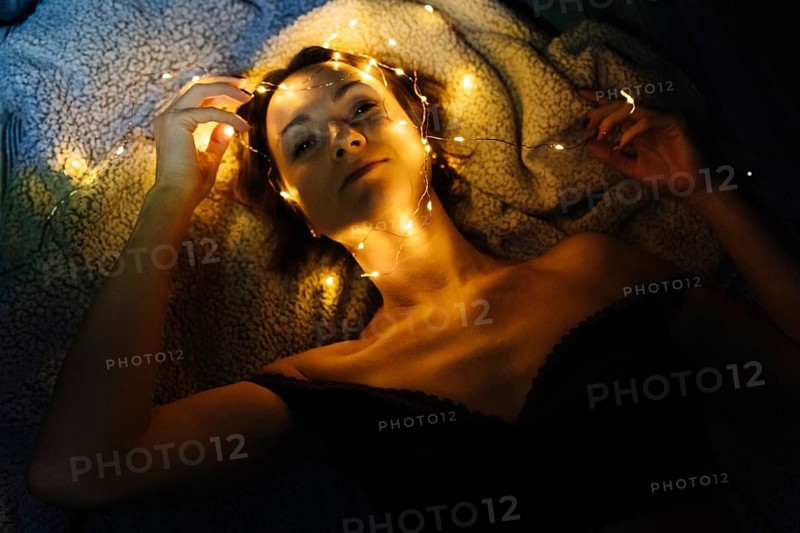 A woman in linen, lowered from her shoulders, lies, decorated with Christmas lights. A woman lies on a textured blanket in a dim environment with warm yellow fairy lights draped around her head, neck and hands. Her relaxed pose and the surrounding darkness emphasize the contrast between the warm illumination and the subdued background, forming an intimate atmospheric portrait with pronounced interplay of light, texture and shadow