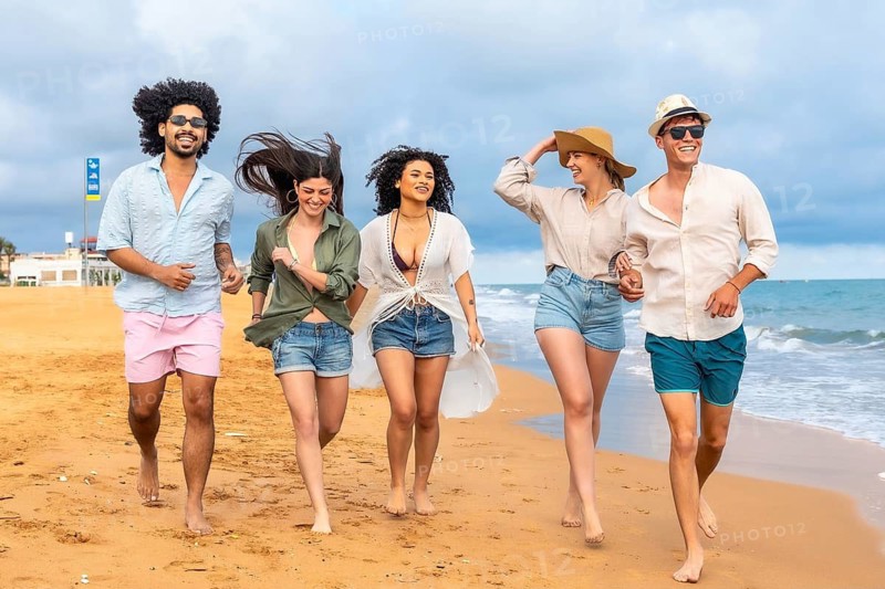 Diverse group of young adult friends enjoying a carefree vacation, running barefoot along the sandy beach with ocean waves gently approaching under a cloudy sky
