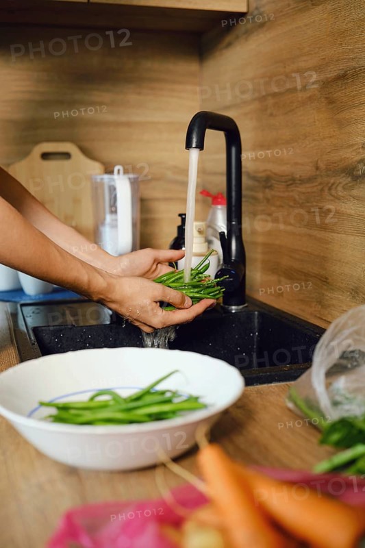 Hands hold green beans under running water in kitchen sink before meal preparation. Bowl with more green beans sits on counter