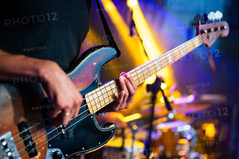 Close-up of a bass guitarist playing during a live concert with colorful stage lighting and blurred drum kit in the background in Lubin, Poland