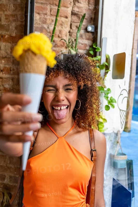 Young woman with curly hair expressing joy and playfulness while holding out a refreshing scoop of sorbet in an ice cream cone on a warm day, enjoying a sweet treat outdoors