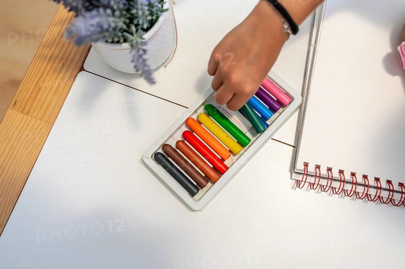 Child's hand selecting wax crayons from a box, preparing to draw on a blank notebook, symbolizing creativity, education, and childhood activities
