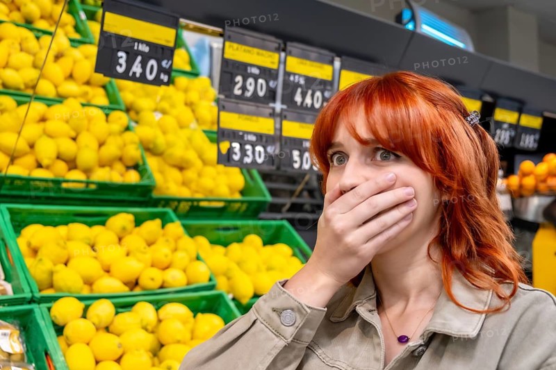 Woman covering her mouth in disbelief and shock while seeing high food prices on price tags in a supermarket, reflecting concerns over inflation and increasing cost of living