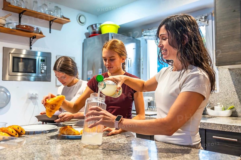 Family members prepare breakfast together in a bright kitchen, pouring juice and arranging croissants, sharing a warm, candid morning moment of bonding and everyday routine
