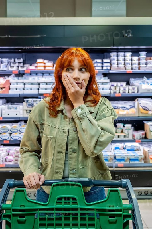 Woman standing in a supermarket aisle with a shopping cart, looking surprised and worried about the increasing cost of food and the economic crisis affecting household budgets