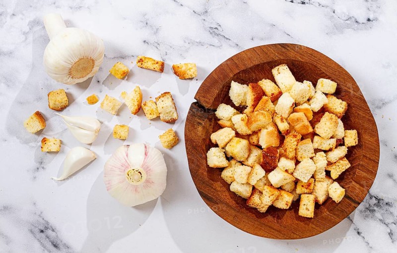 Bread is cut into small pieces and toasted until golden. Garlic sits nearby on a kitchen counter along with a wooden bowl. These croutons are ready for salads or soups