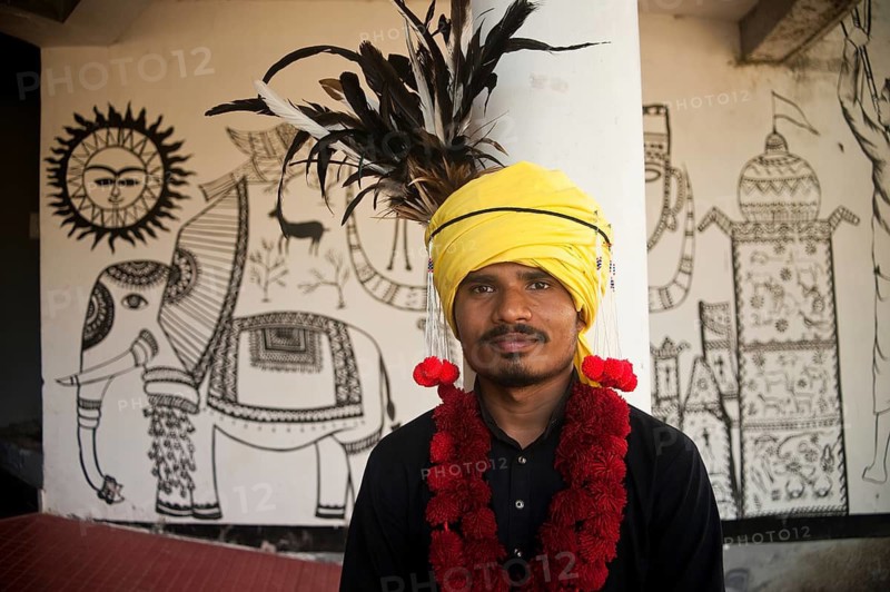 Indian student wearing the Muria tribal men traditional dress, Bastar region, Chhattisgarh, India. In the background, mural paintig inspired by tribal art. Non tribal young man participating at a cultural programme in honour of the Bastar tribal culture