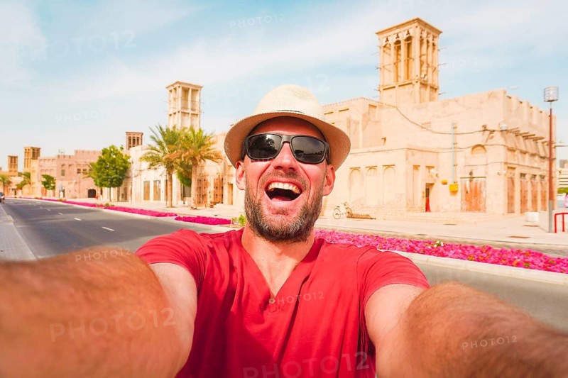 Happy male tourist wear red shirt and straw hat pose takes selfie at the Al Fahidi Historical Neighborhood in Old Dubai. Features a traditional wind tower backdrop cityscape view