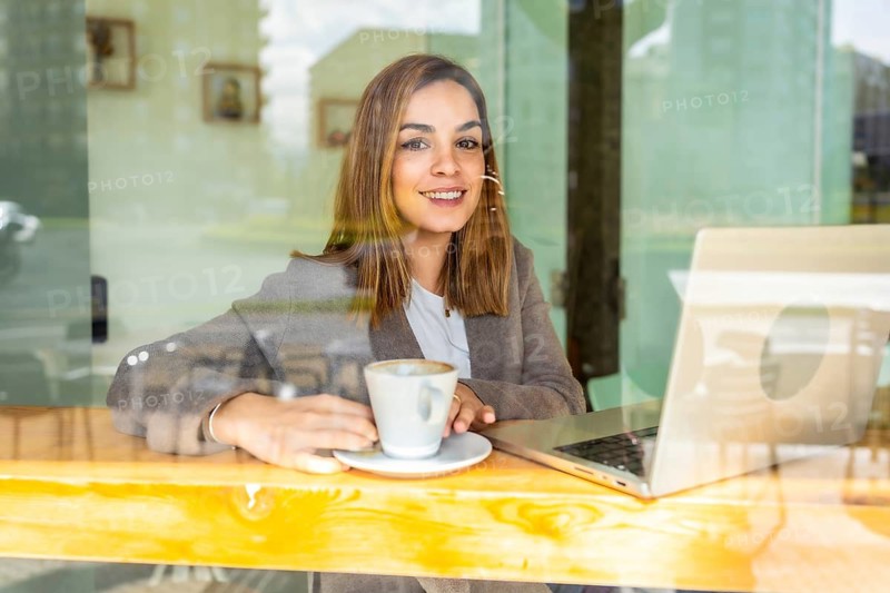 Millennial woman smiling while sitting in a cafe, enjoying a coffee and working on her laptop, reflecting a modern lifestyle of remote work and entrepreneurship