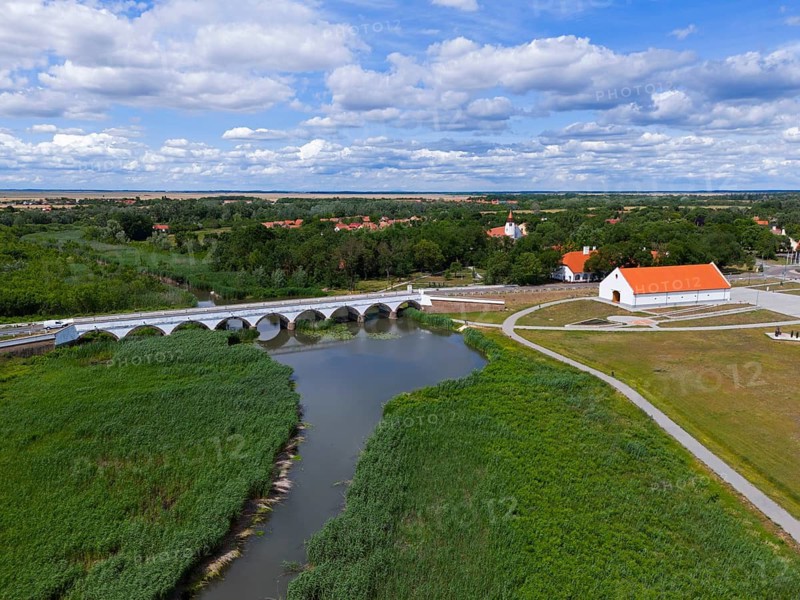Landscape with a river, a bridge and a village surrounded by green meadows under a blue sky with clouds, aerial view, nine-arched stone bridge, Kilenclyukú híd, Hortobagy, Hortobagy National Park, Hortobágyi Nemzeti Park, Hortobágyi Clagoségbolt Park, Puszta, steppe area, UNESCO biosphere reserve, Hungary