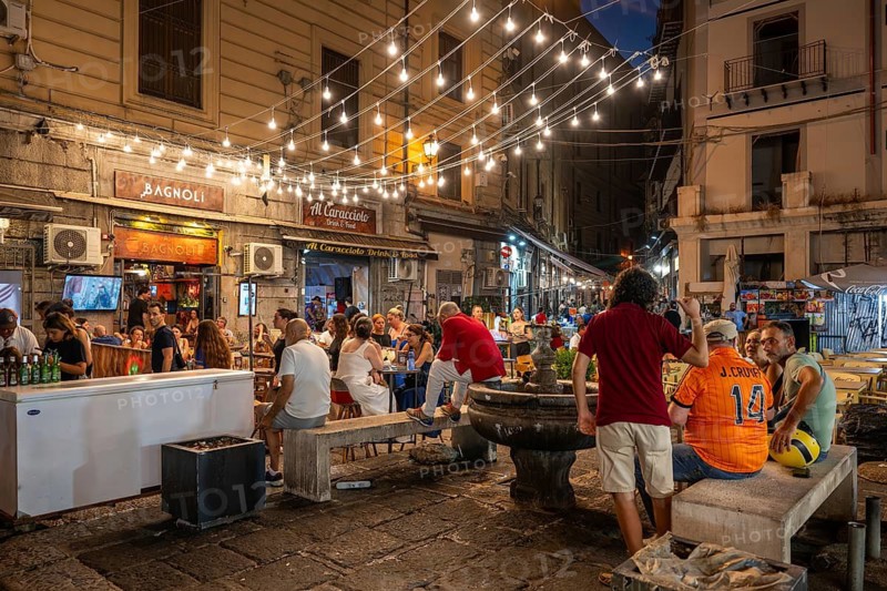 Group of people in a busy square with fountains, fairy lights and restaurants, Palermo, Sicily, Italy