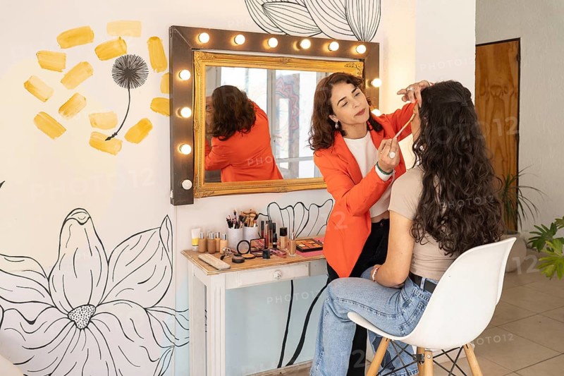 Wide view of a makeup artist applying eye makeup to a seated client at a beauty salon with colorful decor and an illuminated vanity mirror