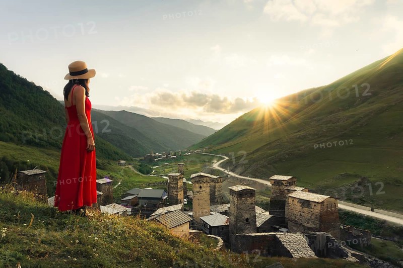 Young pretty Woman traveler in red dress looking at medieval towers of Ushguli Georgia during sunset with mountains and Enguri valley landscape