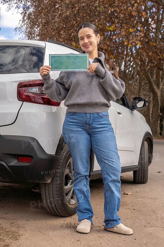 Smiling young woman standing beside white SUV holding blank green sign template, full body shot, outdoor autumn setting