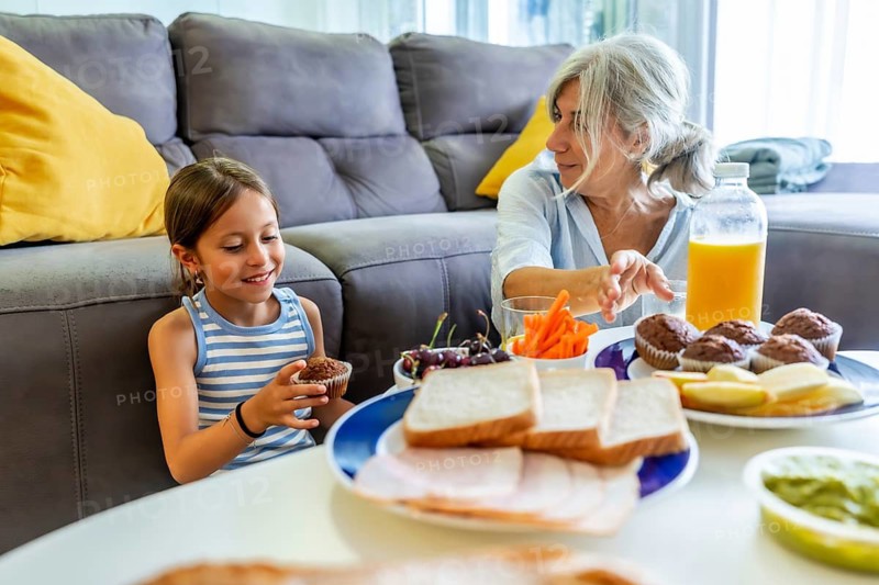 Grandmother and granddaughter are sitting in the living room, enjoying an afternoon snack of muffins, sandwiches, fruit, and juice, sharing a moment of family bonding and care