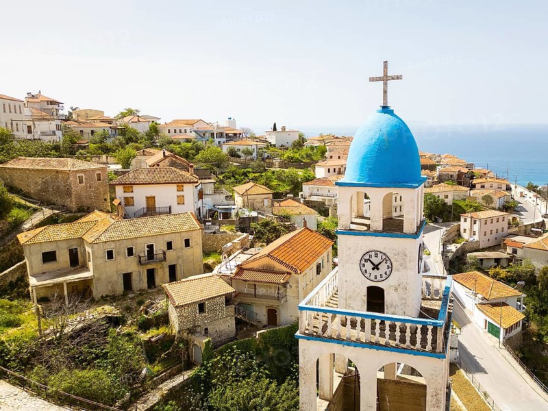 Aerial St. Spyridon Church in Dhrmi. White stone Orthodox chapel with blue domes overlooks Ionian Sea. Historic coastal architecture in Albanian Riviera, Albania