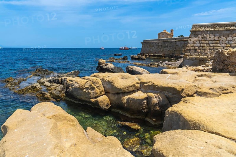 Tabarca island's rocky coastline with clear mediterranean waters, a historic stone wall, and old church building under a summer blue sky, offering a view of boats on the horizon