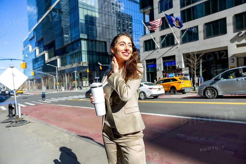 Young business woman smiling, holding a reusable coffee cup, and walking along a sunny city street in manhattan with modern buildings and traffic in the background
