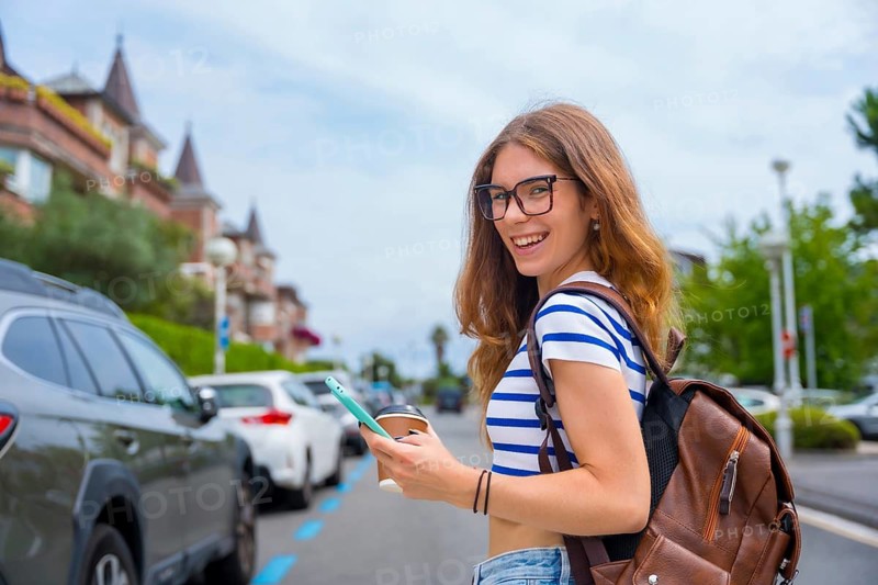 Young woman wearing glasses. A striped top. And a backpack. Walking on a city street while holding a smartphone and a coffee cup