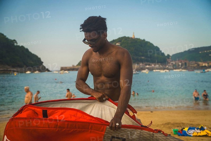 Young shirtless man on a sunny beach opening a vibrant red surf bag, preparing or packing up his paddleboard with the bay and mountainous landscape in the background