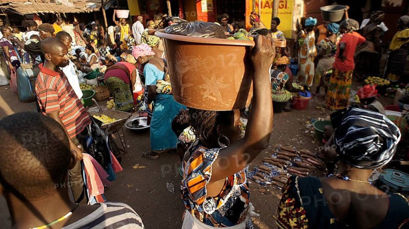 Woman wearing traditional clothes carrying a bucket on her head, lively market with lots of people in colorful clothes carrying goods, Bissau market district, Guinea-Bissau