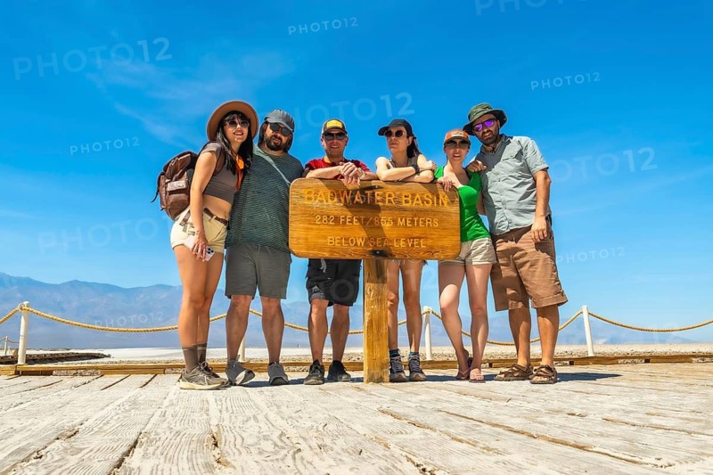 Six friends pose on a wooden boardwalk at badwater basin, smiling at the 282 ft below sea level sign amid vast death valley desert, mountains and bright blue sky