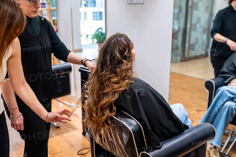 Hairdresser working on a female client's long wavy balayage hair, providing a beauty treatment in a modern hair salon setting, with a second person assisting in the process