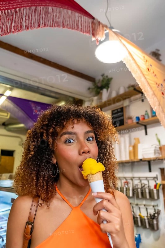 Young woman with curly hair happily eating a scoop of yellow ice cream from a waffle cone, expressing joy and indulgence while standing inside an ice cream parlor