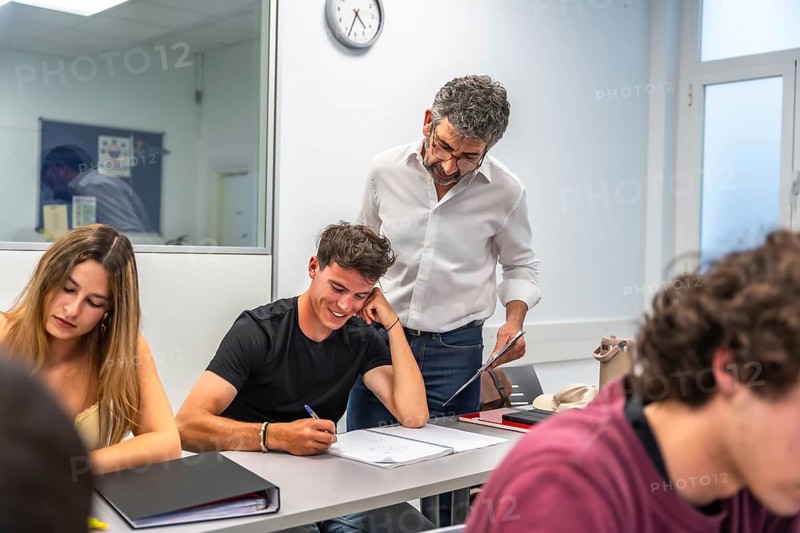 Teacher standing by a male student's desk, assisting him with his work, while other students are focusing on their notes in a bright classroom setting