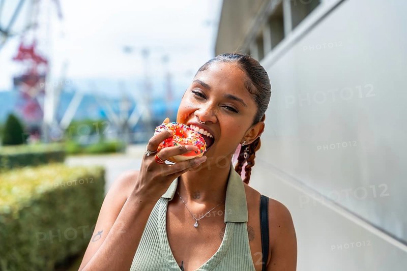 Young woman biting into a colorful glazed donut with sprinkles, expressing happiness and indulgence while enjoying an urban snack outdoors on a sunny day