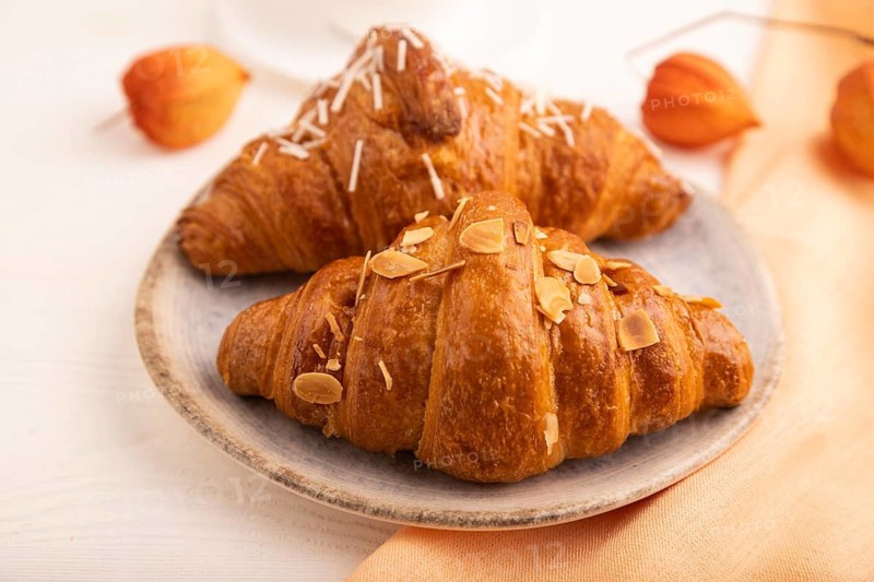 Croissant on blue plate on white wooden background and orange linen textile, cup of coffee, side view, close up