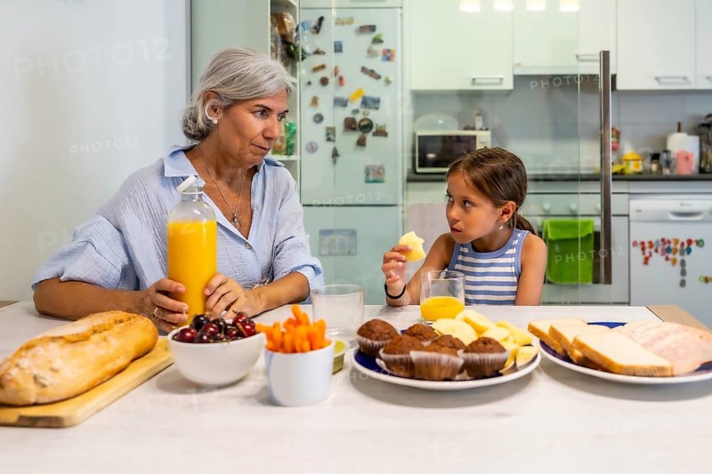 Granddaughter and grandmother enjoying a breakfast together, sitting at the kitchen table while eating and drinking juice, representing a multi generational family bond and care