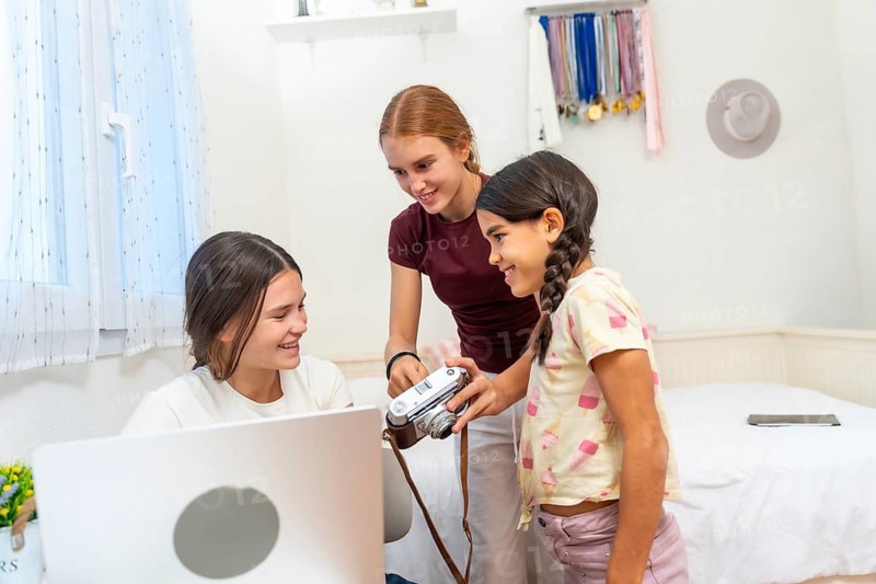 Three sisters laughing and bonding in a bright bedroom as they explore a vintage film camera together with a laptop nearby, sharing curiosity, creativity and everyday joy