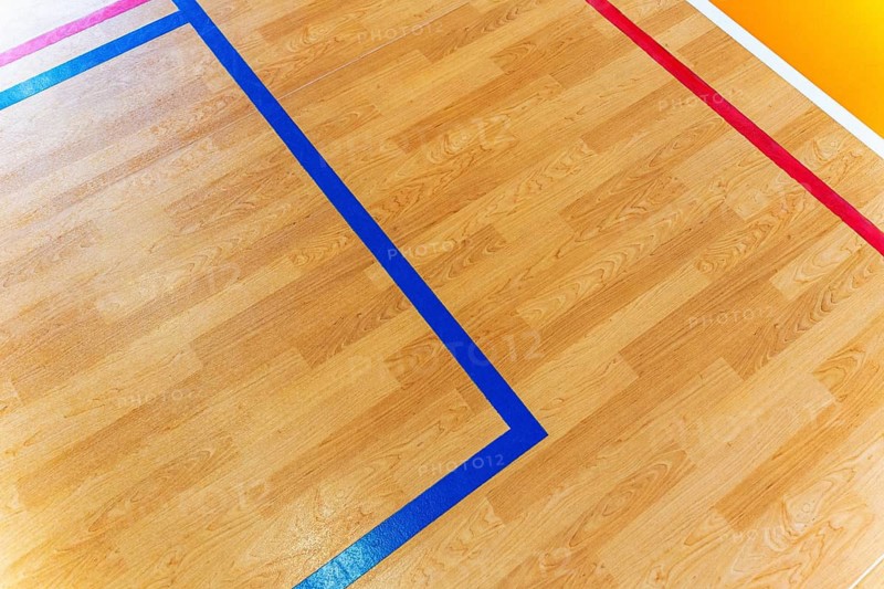 A close-up view of an indoor sports court with blue and red marking lines on a wooden floor. The multicolor layout indicates a multi sport gym used for basketball, volleyball, badminton, handball and other indoor activities. The geometric tape lines create a clear boundary system for various games and training sessions