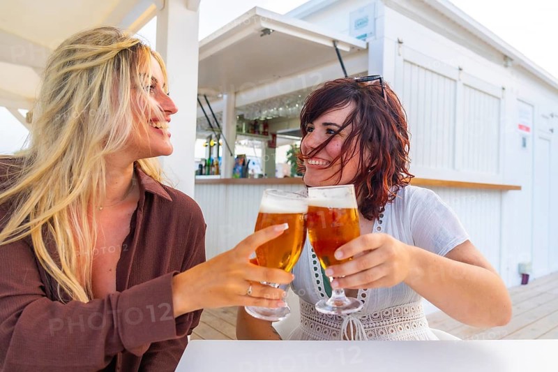 Two happy young women smiling, clinking glasses of refreshing beer, and celebrating friendship outdoors at a beachside bar, reflecting joy and relaxation on a summer day