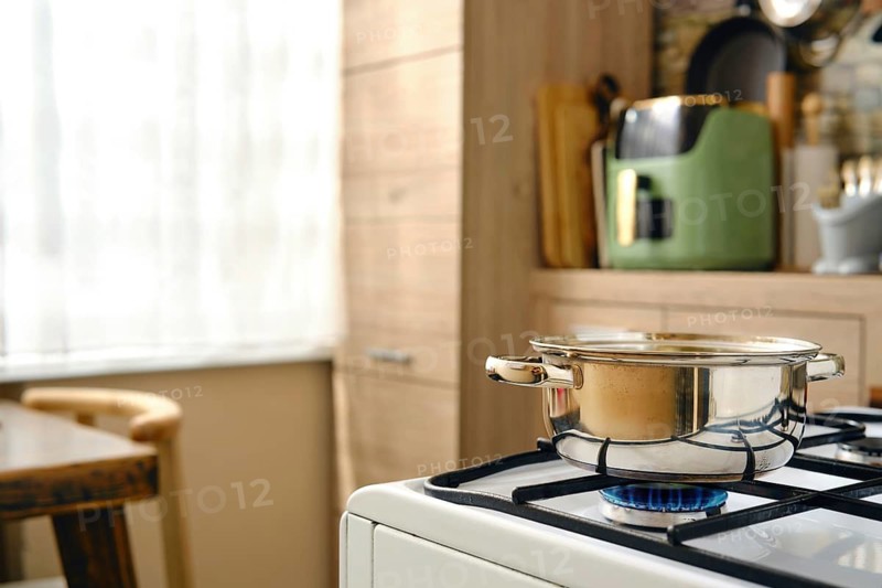 A pot on a gas stove with a blue flame underneath it. Natural light comes through a window in the background. Kitchen cabinets and utensils are visible on the countertop