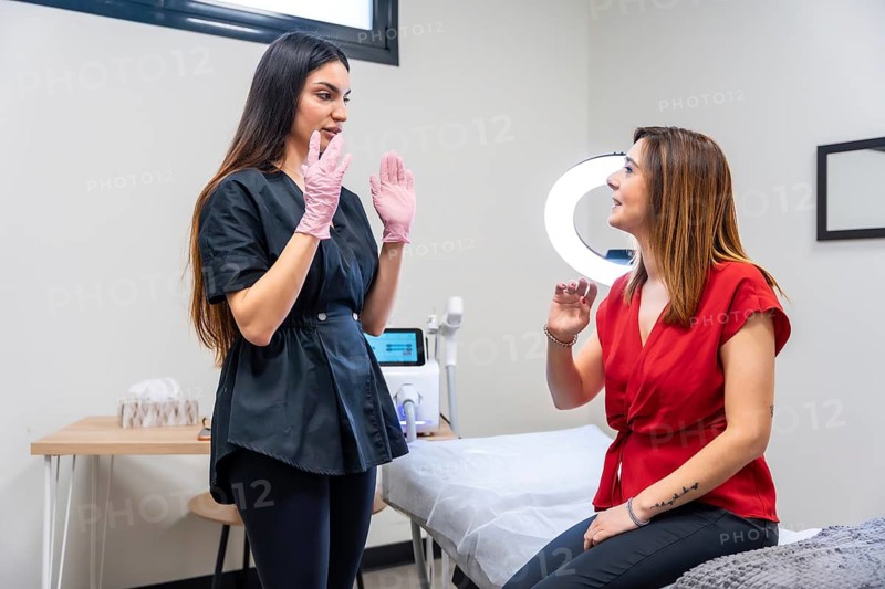 Aesthetic medicine specialist wearing gloves discussing the cosmetic tattoo removal treatment process with a female client sitting on an examination bed in a modern clinic
