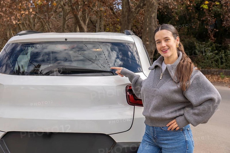 Smiling young woman pointing at rear windshield wiper area of white car, outdoor rural road, car maintenance concept