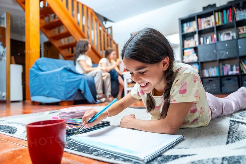 Cheerful preteen girl lying on a patterned rug, smiling and painting with watercolors in a sketchbook while other family members relax in the background