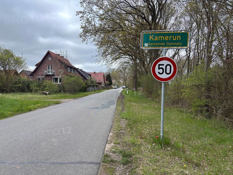 A rural road with speed limit and town sign, surrounded by houses and trees, Cameroon, Wendland, Lower Saxony, Germany