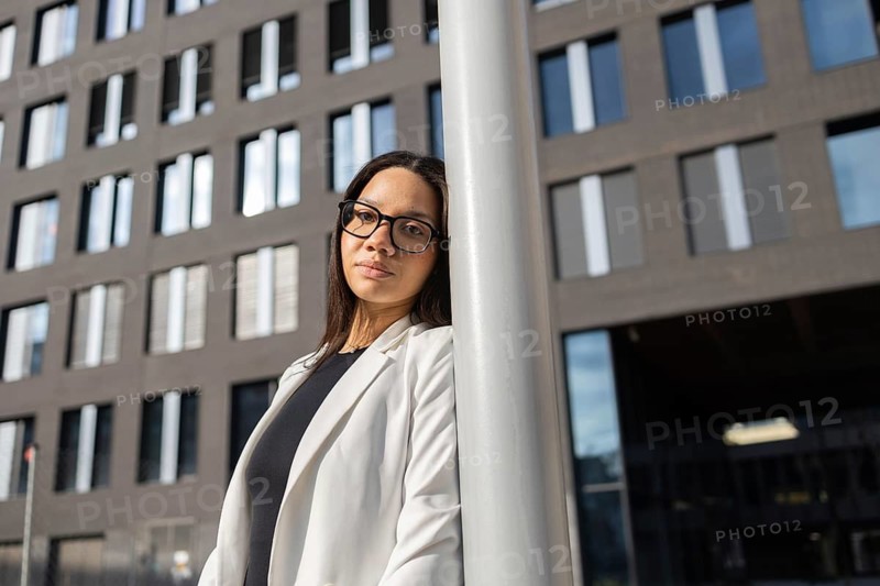 Latin businesswoman in glasses and a white blazer leans on a pole outside a modern office building. Her confident gaze and professional attire reflect success