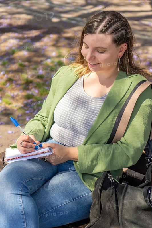 A cheerful young woman with braided hair, captured from a top-down angle, writing in a notebook while sitting on a bench under blooming trees in a peaceful outdoor setting
